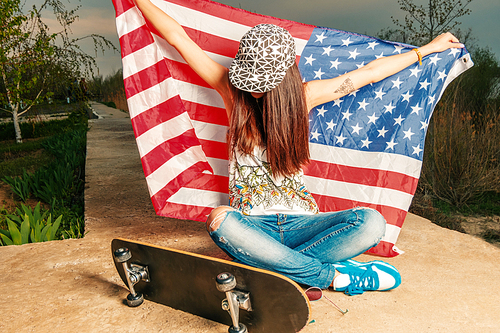 Patriotic girl celebrating legalize with US flag in her hands sitting on lane with her skateboard near