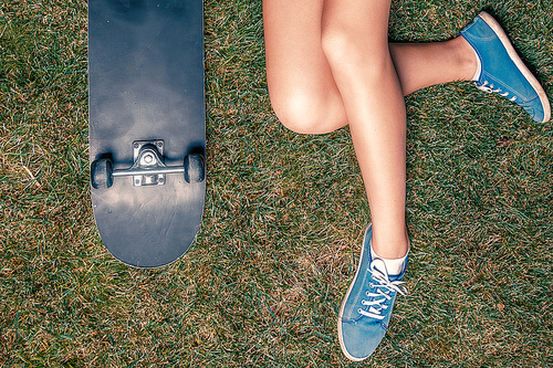 Hipster girl with skateboard resting on green grass in park with her legs crossed
