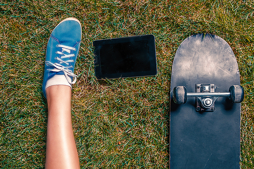 young Girls one leg in teal canvas trainer, tablet computer and skateboard on grass top view