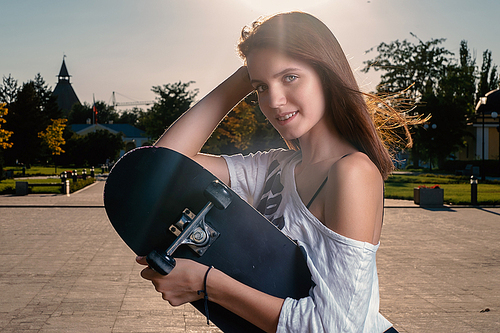 Smiling girl with skateboard in summer city backlit.