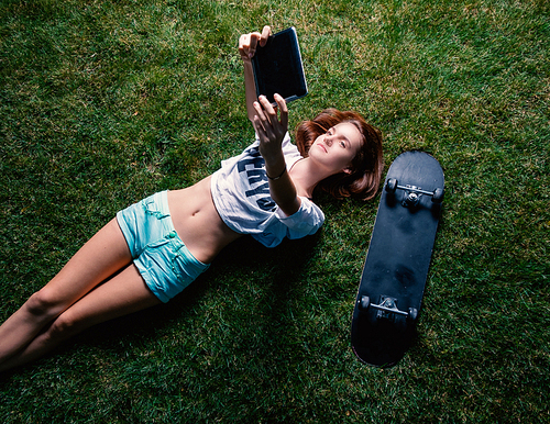 Happy girl lying down the park grass and make selfie with her skateboard lying near top view.