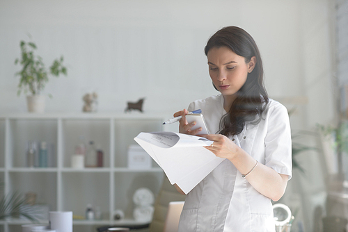 Doctor at her medical office using mobile phone and holding papers. View through the glass