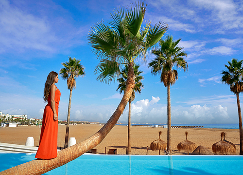 Beautiful woman standing on bent palm tree trunk at infinity pool long red dress