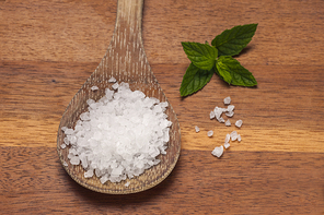 A close up image of a wooden spoon filled with sea salt and a green leaf on a cutting board.