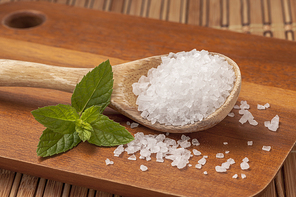 A close up image of a wooden spoon filled with sea salt and a green leaf on a cutting board.