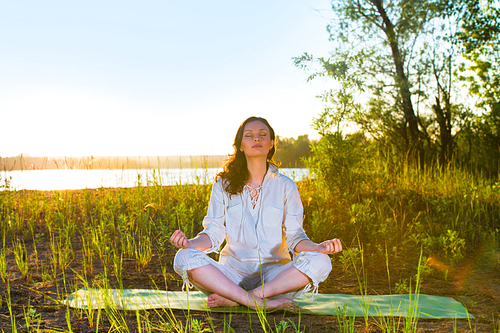 Beautiful young woman doing yoga sitting near the lake early in the morning
