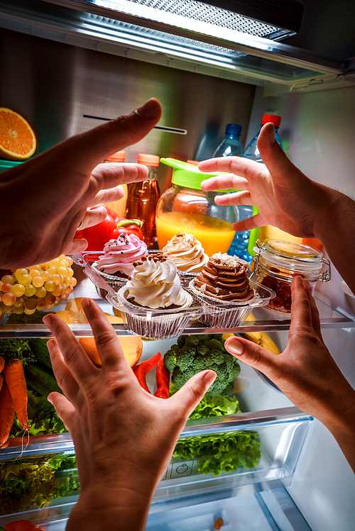 Human hands reaching for sweet cake at night in the open refrigerator