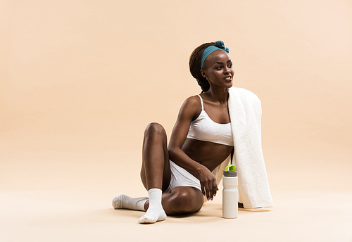 Athletic african girl with a bottle of water and towel sitting on beige studio background