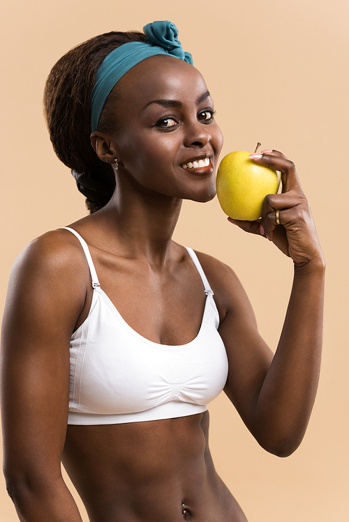 Portrait of sporty african smiling woman eating green apple on beige studio background, 