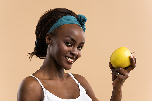 Portrait of sporty african smiling woman eating green apple on beige studio background, 