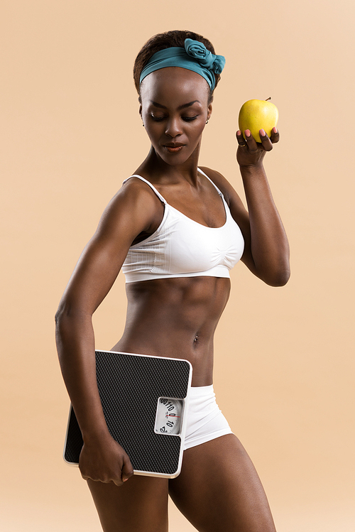 Portrait of attractive african smiling woman eating green apple and holding scales on beige studio background, looking away