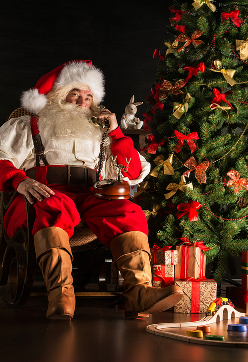 Full length portrait of Santa Claus calling by vintage phone. Sitting near Christmas tree at home