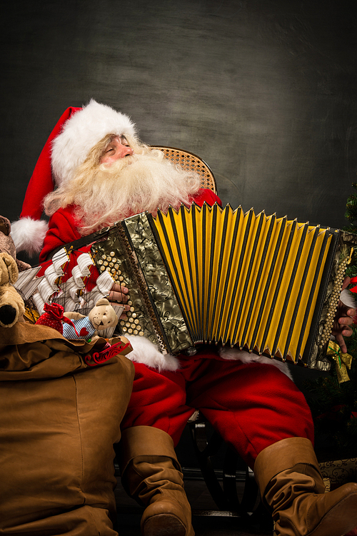 Santa Claus sitting in armchair near Christmas Tree at home and playing music on accordion