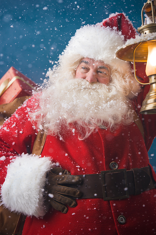 Photo of happy Santa Claus outdoors in snowfall lights the way with vintage lantern while carrying gifts to children