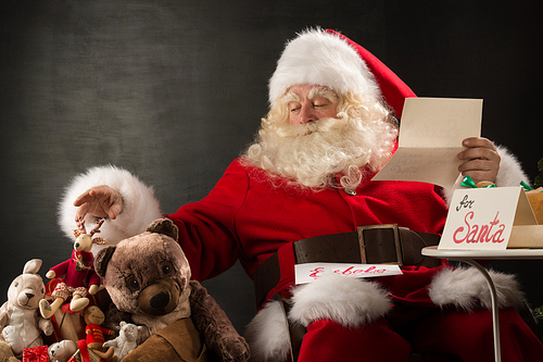 Santa Claus sitting at home at comfortable armchair holding envelope and reading children's letters and wishes and choosing toys from big sack near him.