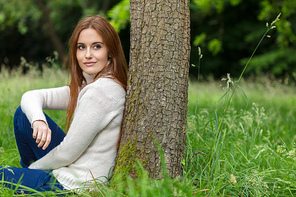Outdoor portrait of beautiful thoughtful happy girl or young woman with red hair wearing a white jumper sitting & leaning against a tree in the countryside