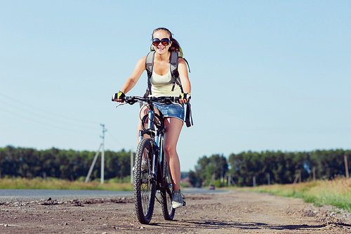 Young sporty girl with backpack riding bicycle along roadside