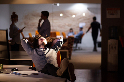 Full length of a relaxed casual young businessman sitting with legs on desk at night office