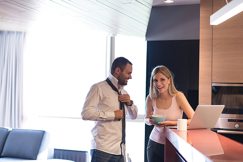 A young couple is preparing for the job and using a laptop. The man drinks coffee while the woman eats breakfast at luxury home together, looking at screen, smiling.