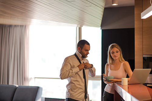 A young couple is preparing for the job and using a laptop. The man drinks coffee while the woman eats breakfast at luxury home together, looking at screen, smiling.