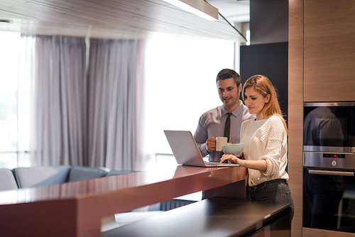 A young couple is preparing for the job and using a laptop. The man drinks coffee while the woman eats breakfast at luxury home together, looking at screen, smiling.