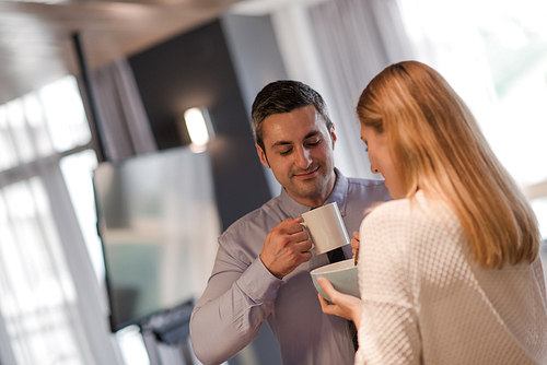 A young couple is preparing for the job and using a laptop. The man drinks coffee while the woman eats breakfast at luxury home together, looking at screen, smiling.