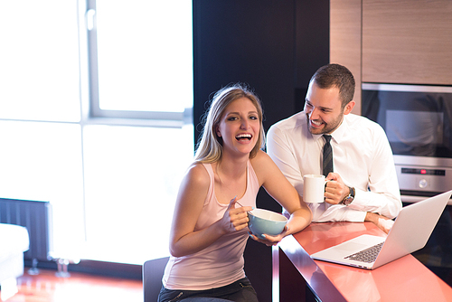 A young couple is preparing for the job and using a laptop. The man drinks coffee while the woman eats breakfast at luxury home together, looking at screen, smiling.