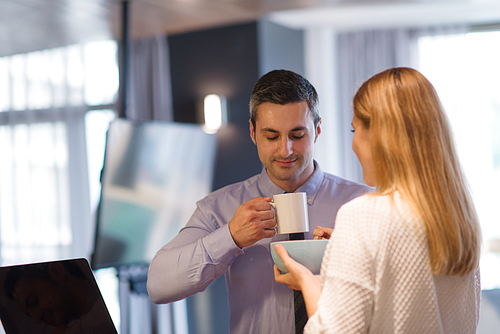 A young couple is preparing for the job and using a laptop. The man drinks coffee while the woman eats breakfast at luxury home together, looking at screen, smiling.