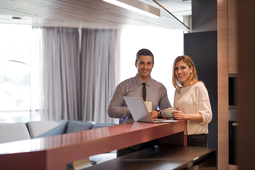 A young couple is preparing for the job and using a laptop. The man drinks coffee while the woman eats breakfast at luxury home together, looking at screen, smiling.