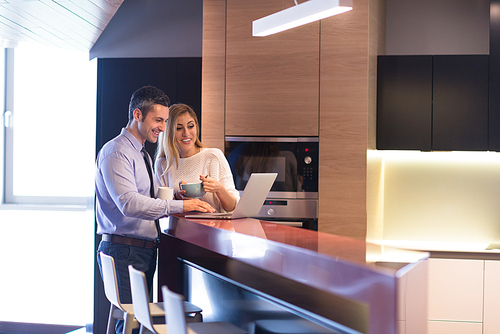 A young couple is preparing for the job and using a laptop. The man drinks coffee while the woman eats breakfast at luxury home together, looking at screen, smiling.