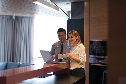 A young couple is preparing for the job and using a laptop. The man drinks coffee while the woman eats breakfast at luxury home together, looking at screen, smiling.