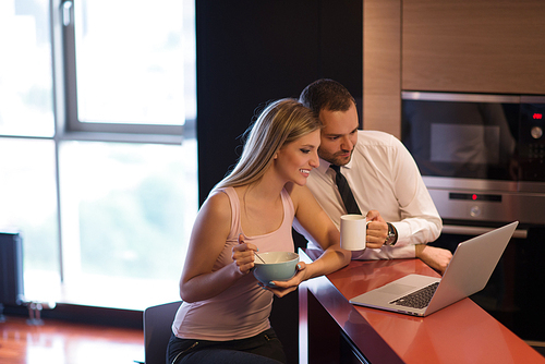 A young couple is preparing for the job and using a laptop. The man drinks coffee while the woman eats breakfast at luxury home together, looking at screen, smiling.