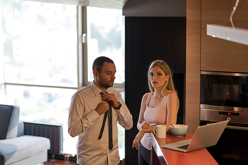 A young couple is preparing for the job and using a laptop. The man drinks coffee while the woman eats breakfast at luxury home together, looking at screen, smiling.