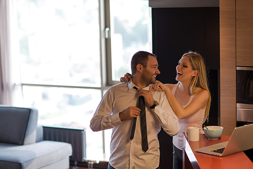 A young couple is preparing for the job and using a laptop. The man drinks coffee while the woman eats breakfast at luxury home together, looking at screen, smiling.