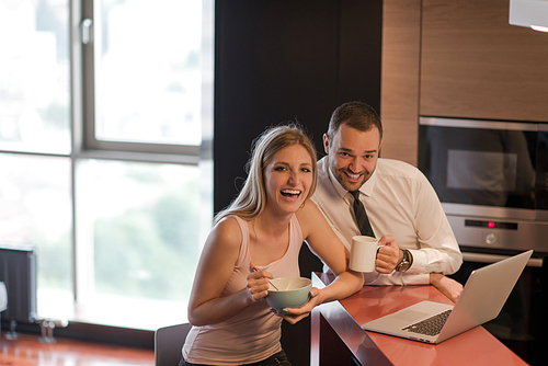 A young couple is preparing for the job and using a laptop. The man drinks coffee while the woman eats breakfast at luxury home together, looking at screen, smiling.