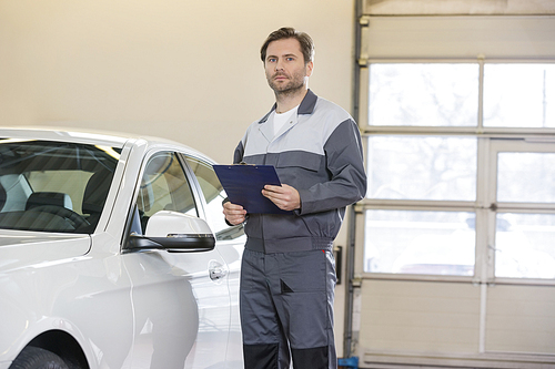 Portrait of confident male mechanic with clipboard standing by car in workshop