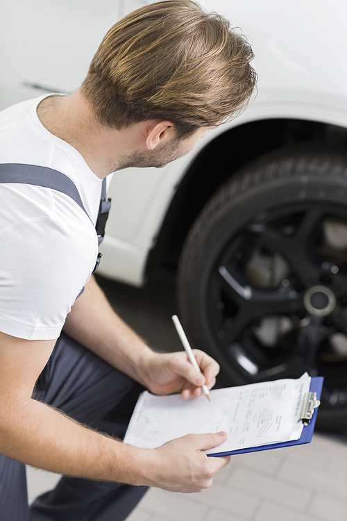 Side view of automobile mechanic writing on clipboard while examining car's wheel in workshop