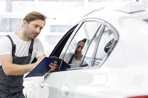 Automobile mechanic writing on clipboard while examining car in repair shop