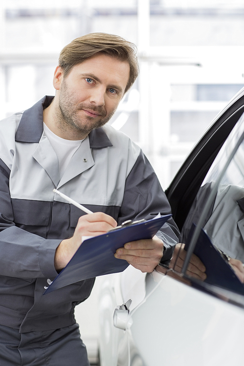 Portrait of confident mechanic holding clipboard while leaning on car's window in workshop