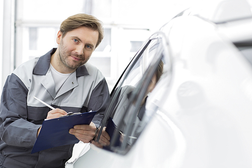 Portrait of confident automobile mechanic holding clipboard while leaning on car's window in workshop