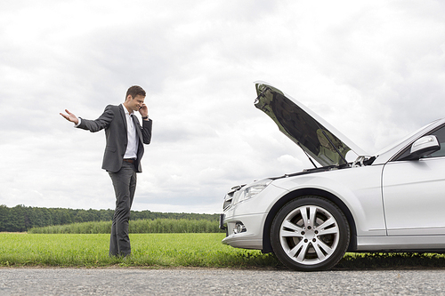 Frustrated young businessman using cell phone by broken-down car at countryside