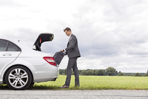 Full length side view of young businessman unloading luggage from broken down car at countryside