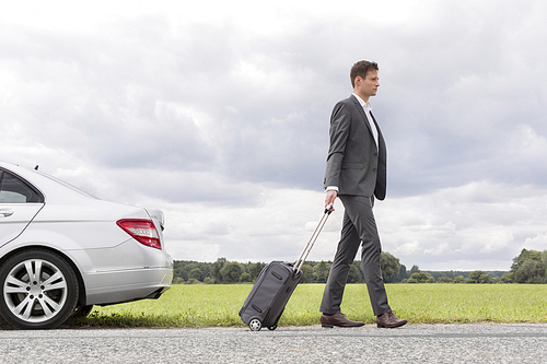 Full length side view of young businessman with suitcase leaving broken down car at countryside