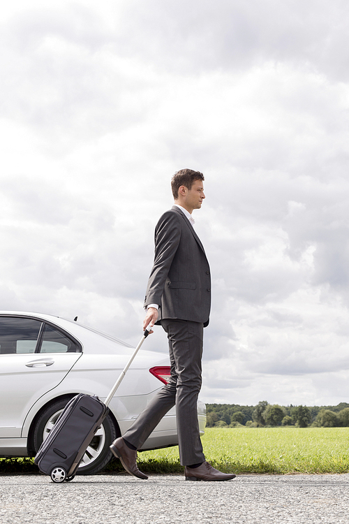 Full length side view of young businessman with luggage walking by broken down car at countryside