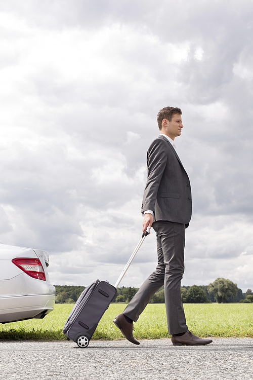 Full length side view of young businessman with luggage leaving broken down car at countryside