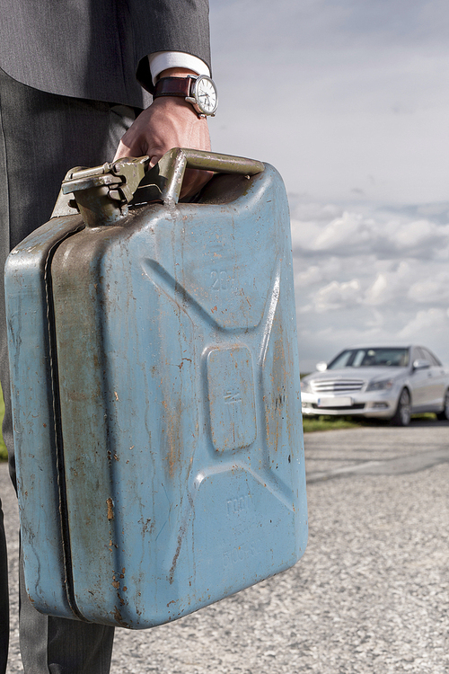 Low section of young businessman carrying gasoline can with broken car in background at countryside