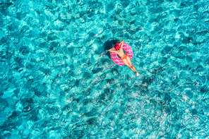 Aerial view of slim young woman swimming on the pink donut swim ring in the transparent blue sea at sunny bright day. Tropical aerial landscape with girl, azure water. Top view. Summer travel. Holiday
