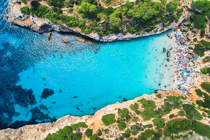 Aerial view of sandy beach with colorful umbrellas, swimming people in sea with transparent blue water and green trees on the mountain at sunrise in summer. Travel in Mallorca, Spain. Top view. Nature
