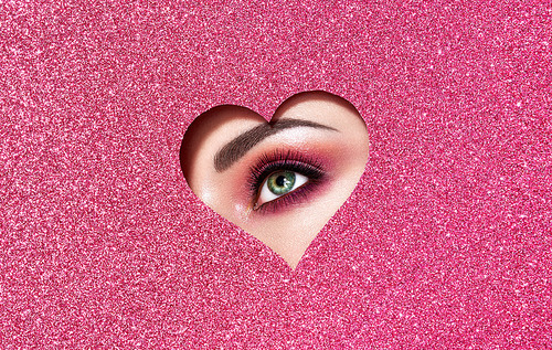 Conceptual photo of Valentine's day. Eye of Girl with Festive Pink Makeup. Paper heart on a pink background. Love symbols Valentines day
