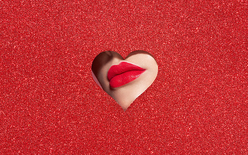 Beautiful Plump Bright Lips Of a Young Beautiful Woman with Red Lipstick Look Into the Pattern of Heart Shaped made of Colored Paper. Holiday Patterns. Valentine's Day. Beautiful Love Make-up